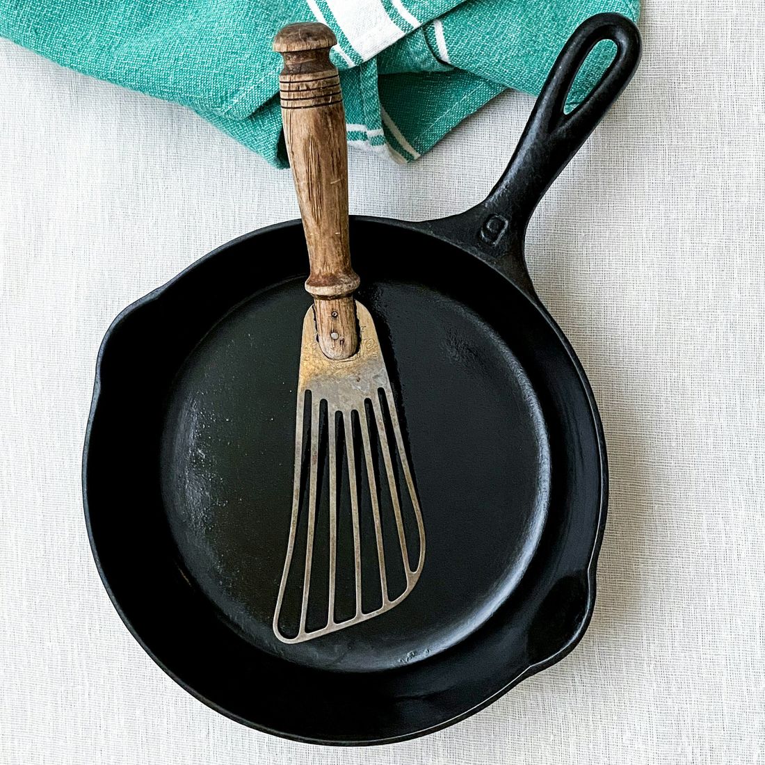 An empty black cast iron skillet seen from above with a metal spatula and a green kitchen towel on a light linen cloth.