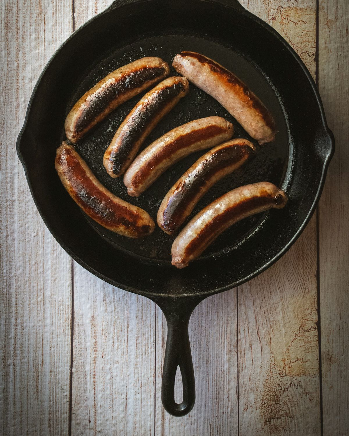 Top view of a black cast iron skillet with six browned sausages, on a light wooden table.